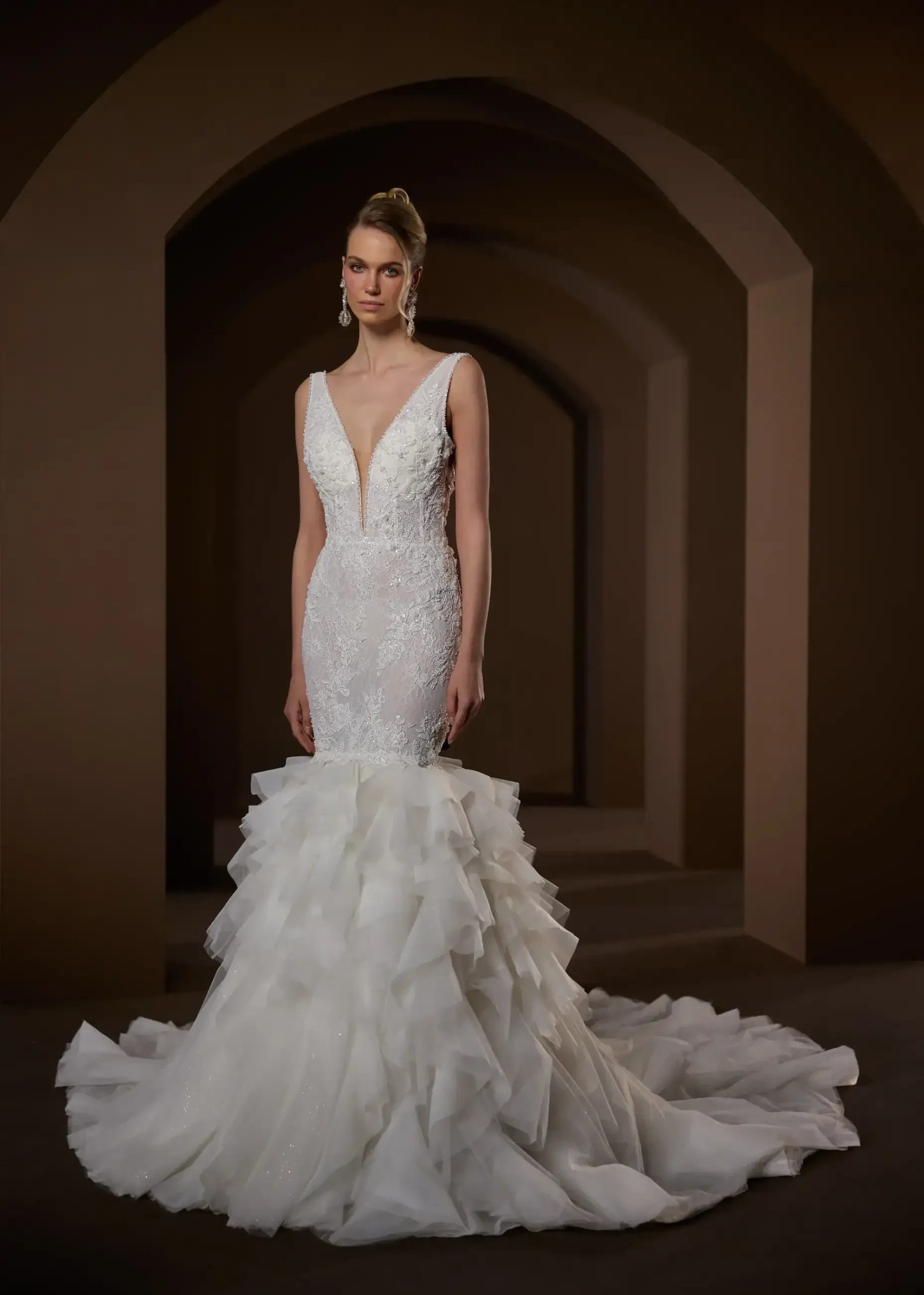 Model is wearing a sleeveless bridal lace mini dress with an optional detachable skirt that transforms it into a mermaid dress with full layers of tulle and a long train.