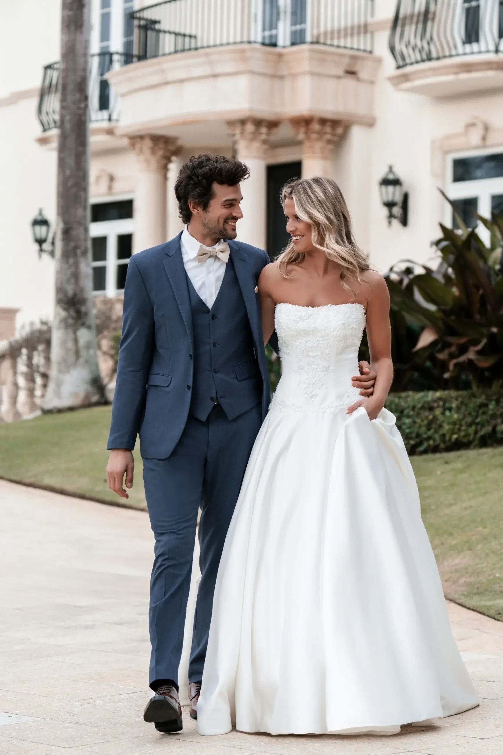 A couple dressed for a wedding, the man in a blue suit and the woman in a white strapless wedding gown, walking together and smiling.