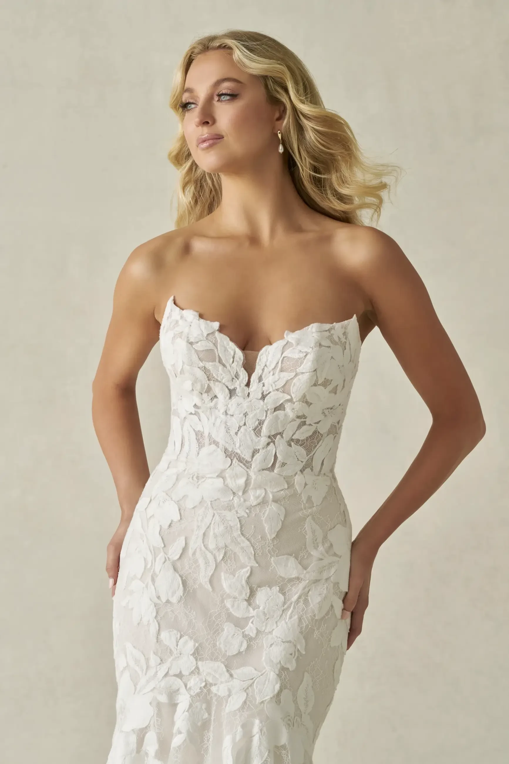 A model wearing a strapless white wedding dress with floral lace detailing, standing elegantly with long wavy hair.