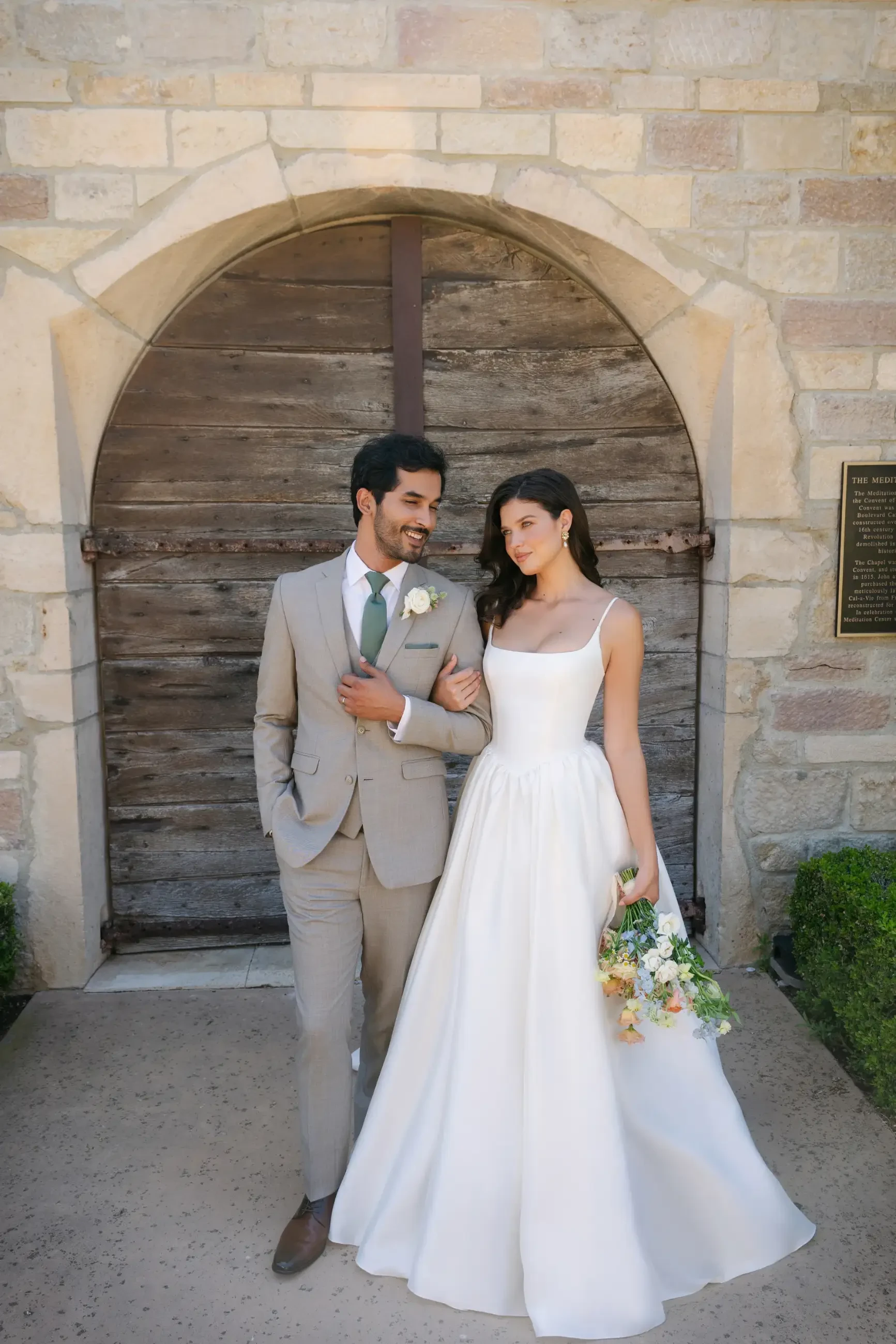 A bride and groom stand arm-in-arm, smiling, in front of a rustic wooden door. The bride wears a white gown and holds a bouquet, radiating joy.