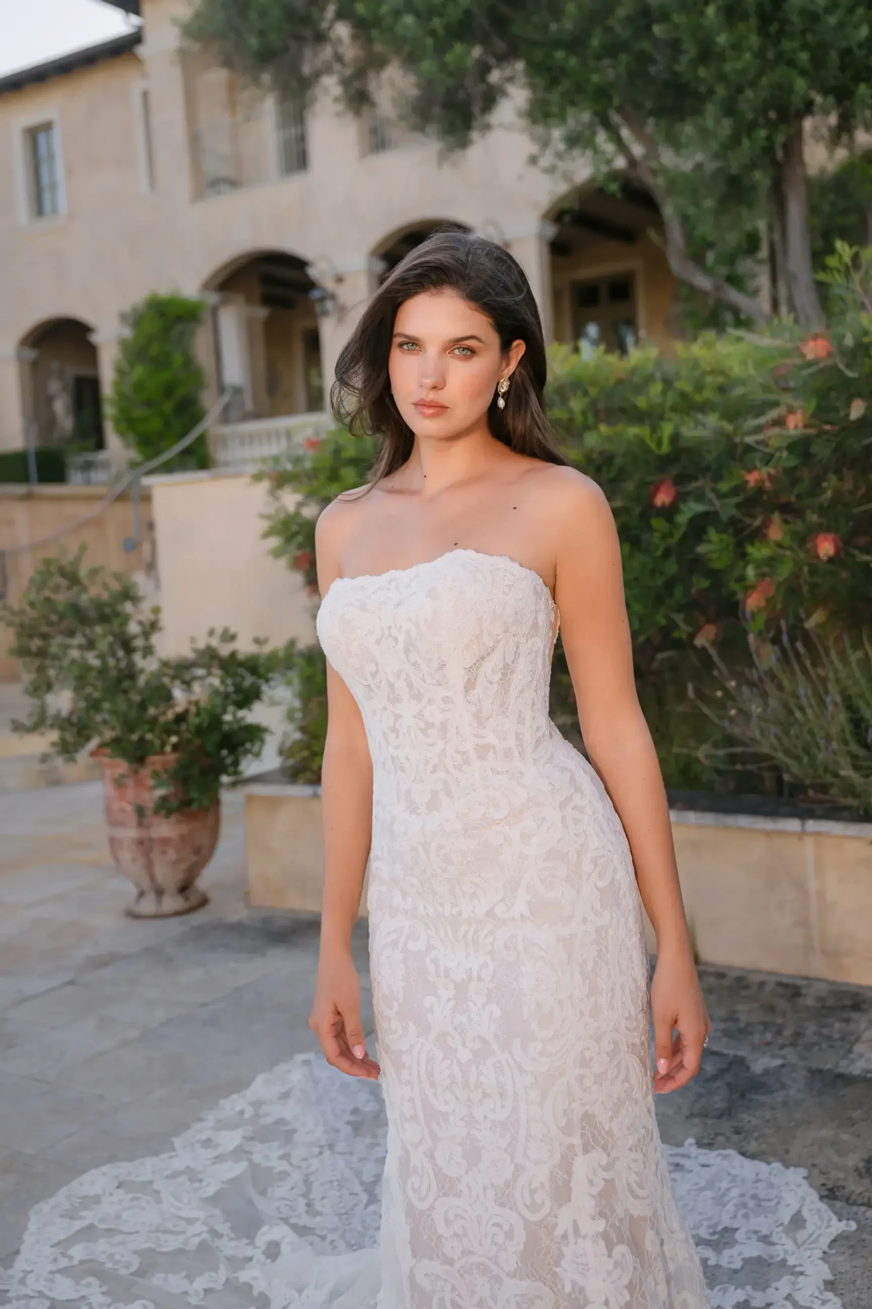 A woman in a strapless lace wedding gown stands in an elegant courtyard. She looks confidently at the camera, with greenery and a grand building behind her.