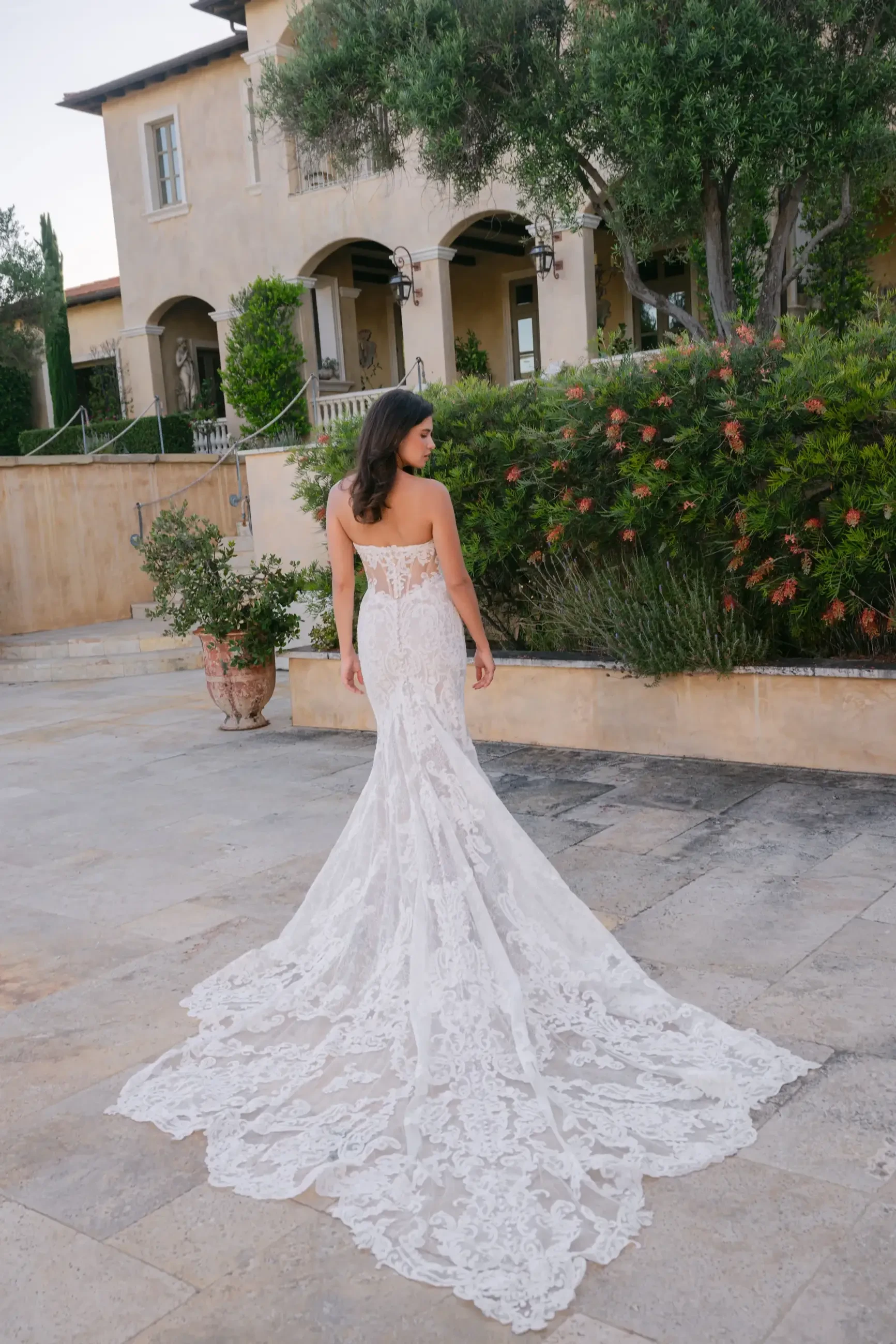 A woman in a strapless lace wedding gown stands with her back to the camera, showcasing the long train of the dress. She is outside, near a building and surrounded by greenery.