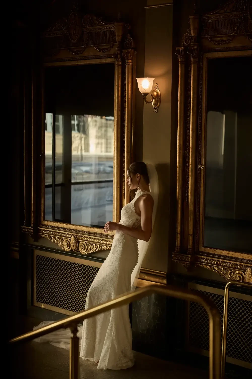 A bride in a white wedding dress leans against a wall next to a large mirror, showcasing elegant decor and soft lighting.