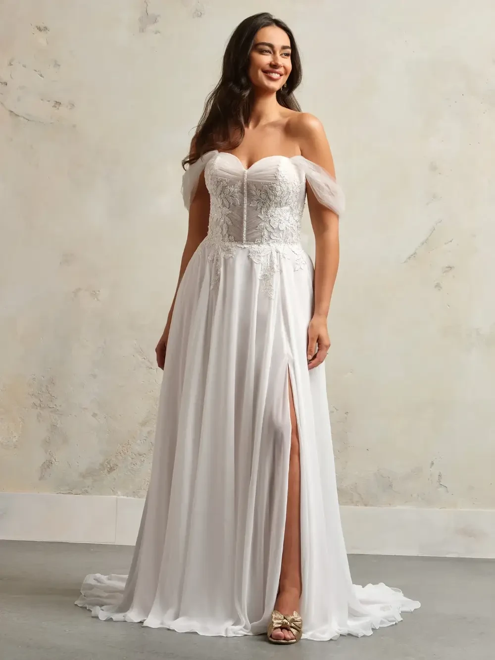 A woman wearing a white, off-the-shoulder wedding dress with a slit and intricate lace detailing, standing against a neutral background.