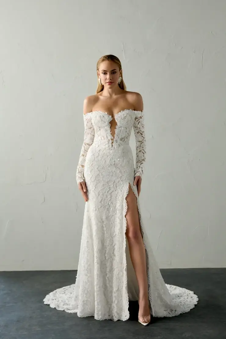 A model wearing an elegant, off-the-shoulder white lace gown with long sleeves and a high slit, standing against a plain background.