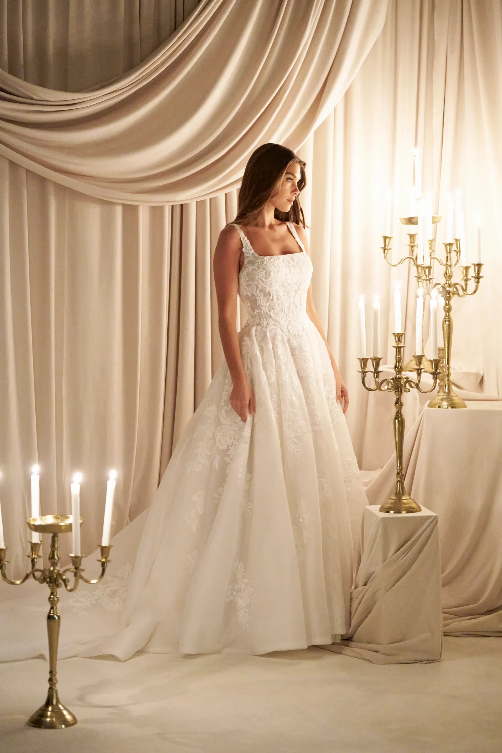 A woman in an intricately beaded white gown holds a lit candle. She stands amid brass candelabras and draped curtains, evoking an elegant, serene ambiance.