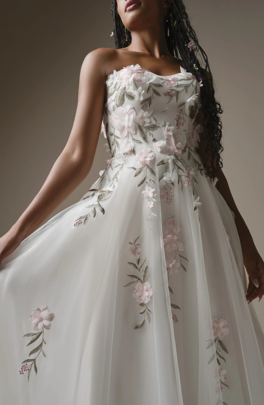 A woman in a delicate, strapless white gown with pink floral embroidery holds the flowing dress. The mood is elegant and serene.