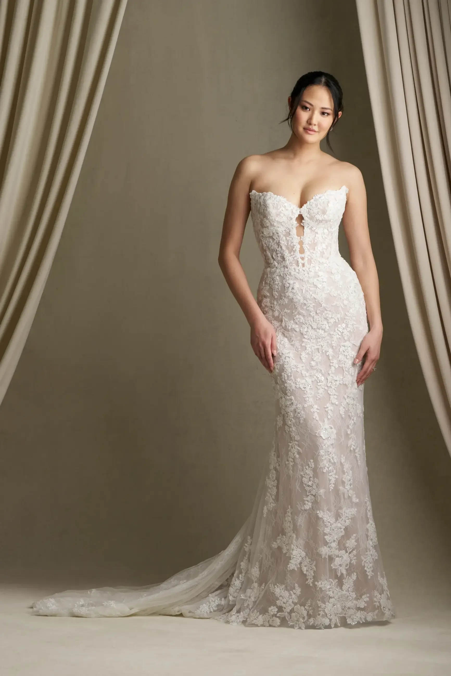 A woman in an elegant, strapless lace wedding gown with a subtle train stands gracefully against a neutral draped curtain backdrop, exuding a tranquil, sophisticated aura.