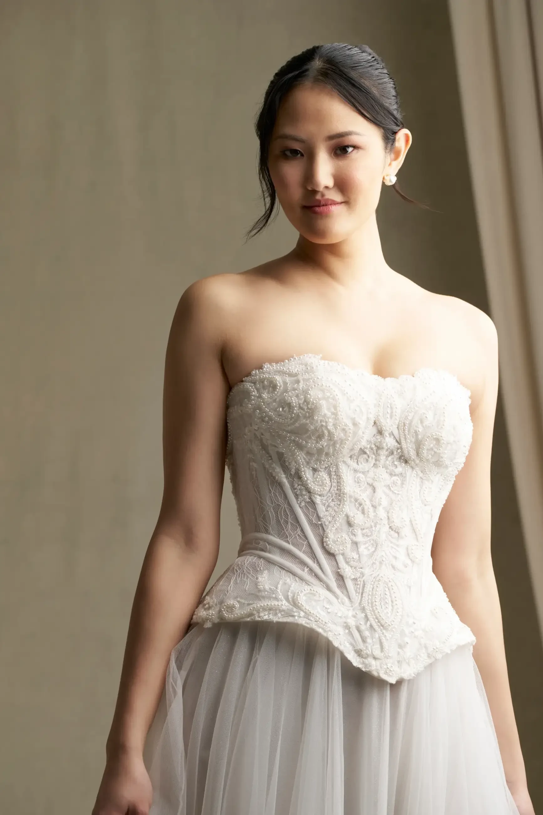 A woman in a strapless, intricately beaded white wedding dress stands against a soft beige background, exuding elegance and confidence.