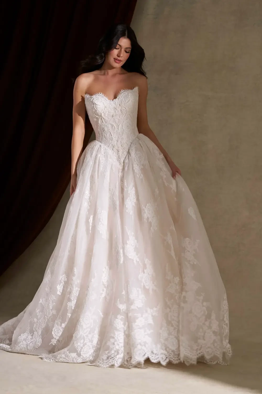 A woman in a strapless, layered white wedding dress with intricate lace detailing, standing gracefully against a neutral background.