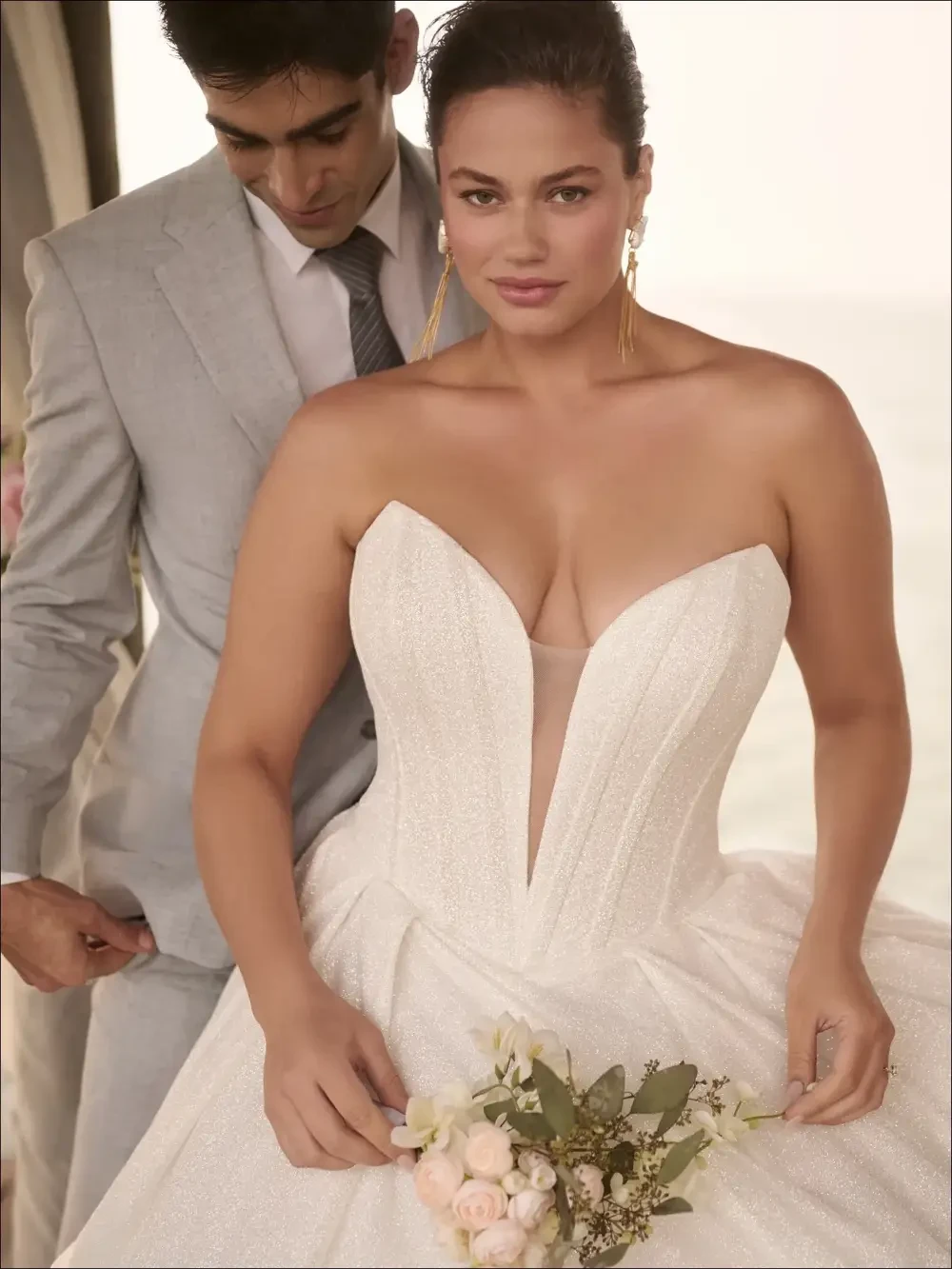 A bride in a strapless wedding dress with a plunging neckline holds a bouquet, while a man in a light gray suit stands behind her.