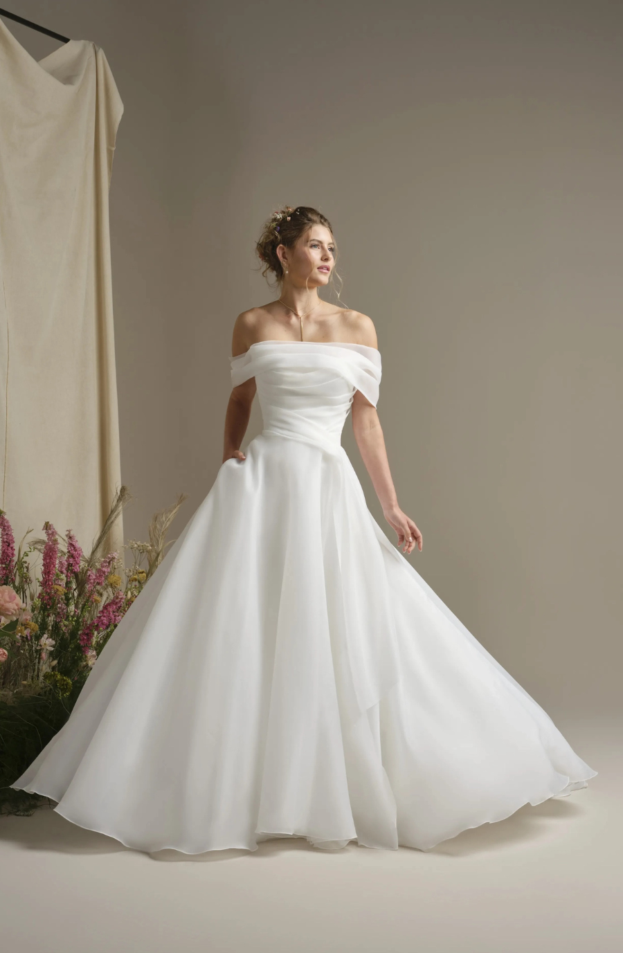 A woman in an elegant off-shoulder white wedding dress stands gracefully, surrounded by flowers. The scene conveys a serene, romantic atmosphere.