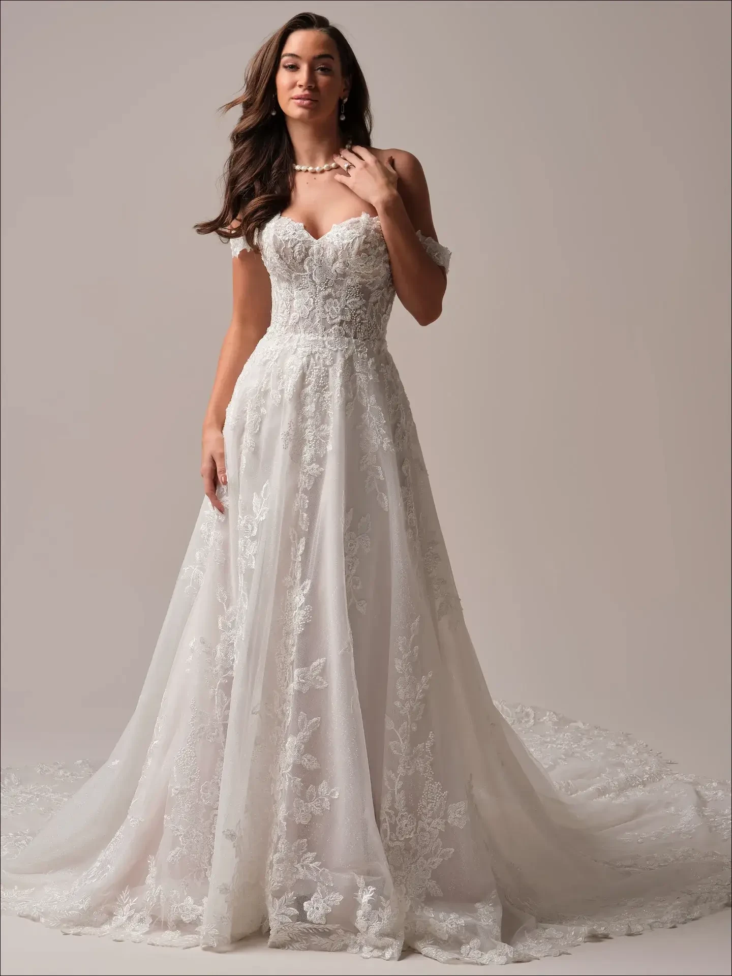 Model is wearing an off-the-shoulder A-line wedding gown with lace embellishments and a romantic flowing skirt.