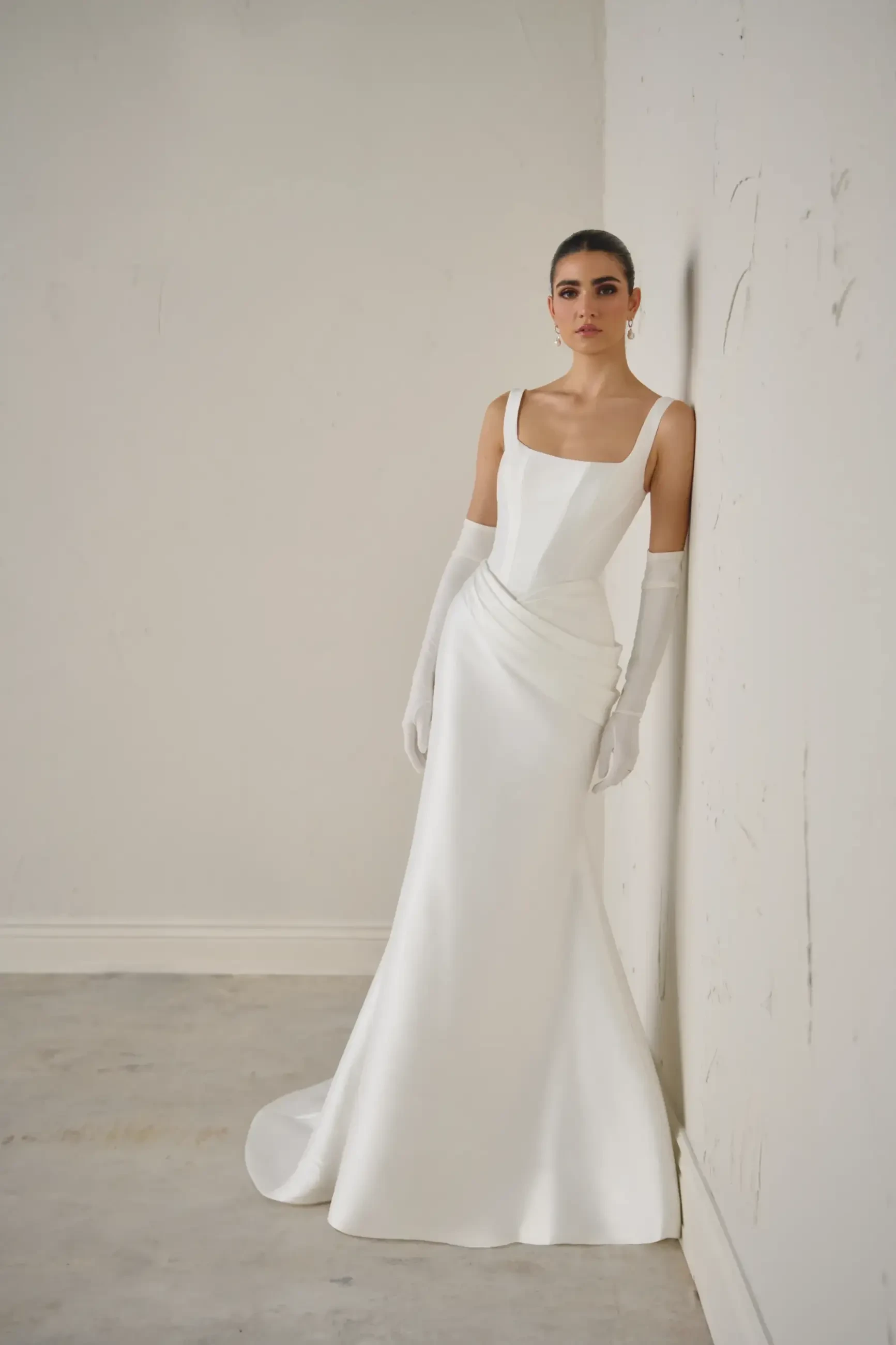 Elegant woman in a sleek white wedding gown with a square neckline and long gloves, leaning against a minimalist white wall. Timeless and sophisticated atmosphere.