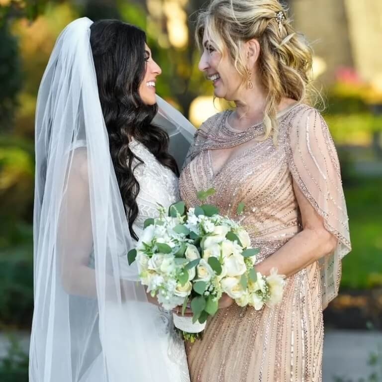 A bride in a white gown and veil smiles at a woman