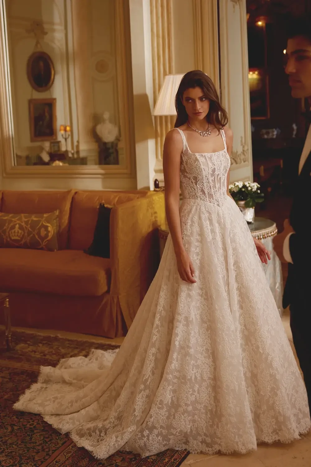 A model wearing an elegant, lace wedding dress stands in a luxurious room with a sofa and decorative items.