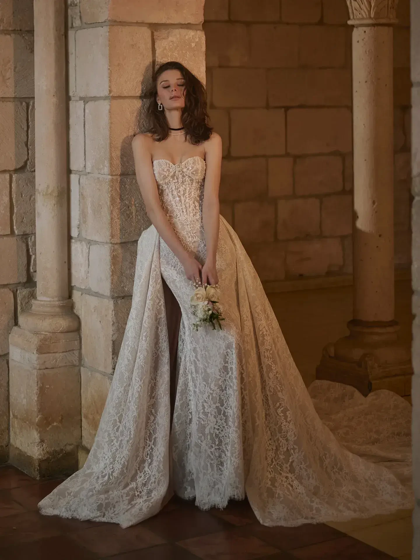 Model is wearing a Chantilly lace mermaid bridal dress with an illusion back and an optional overskirt.