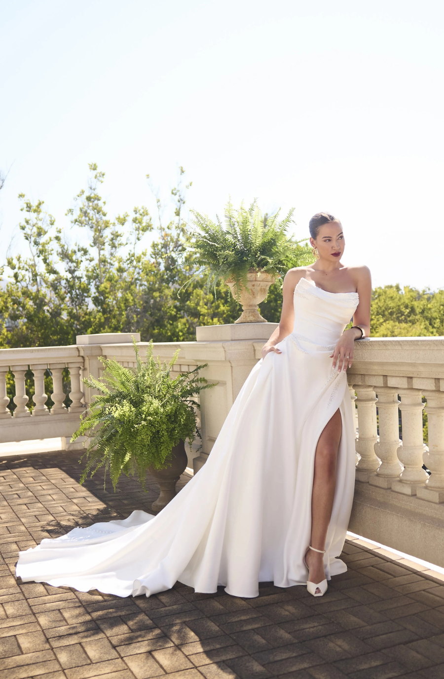 A woman in a strapless white gown stands on a terrace with lush greenery. She exudes elegance with a confident pose and serene expression under a clear sky.