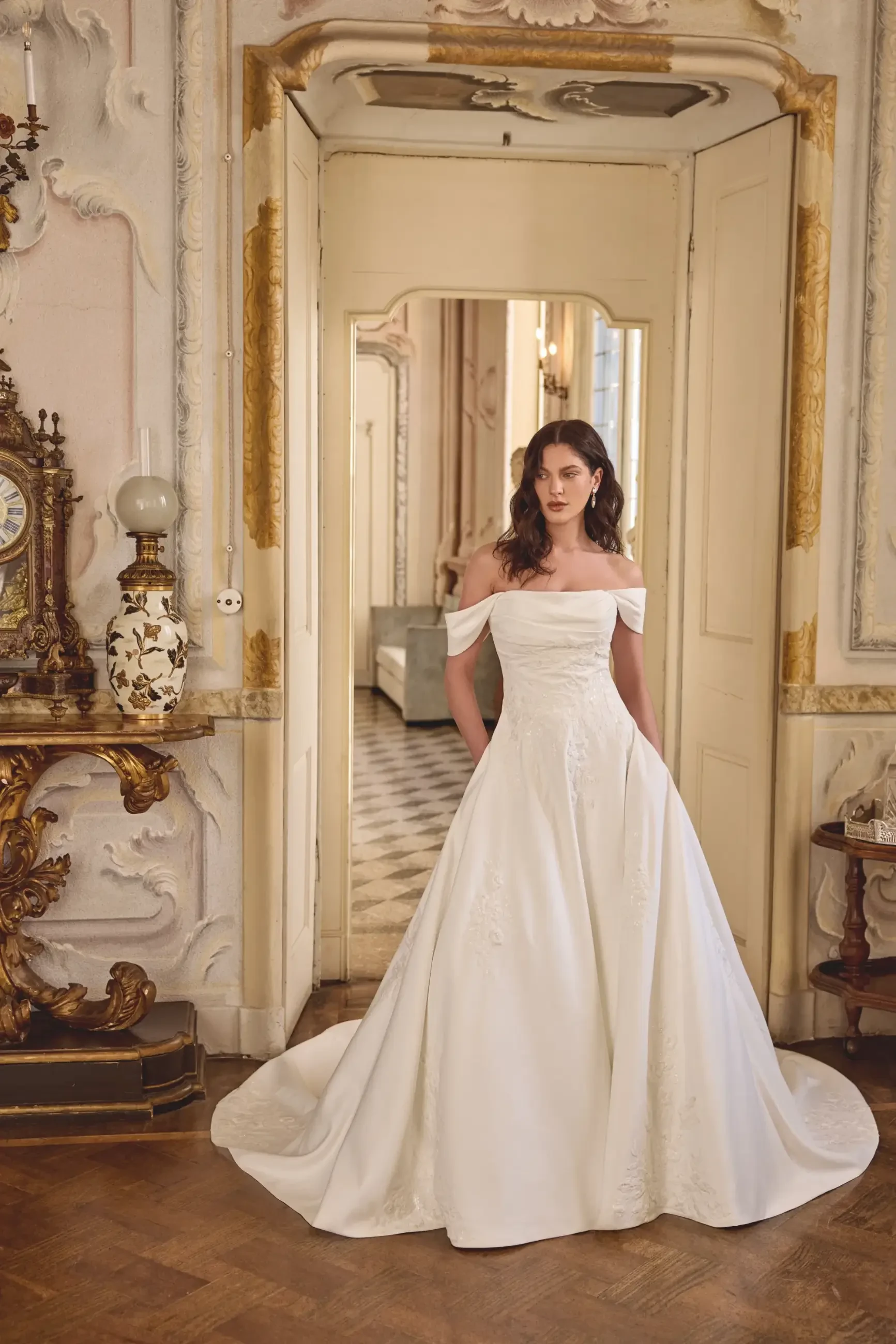 A model wearing a white, off-the-shoulder wedding gown stands in an elegant room with ornate decorations and a mirror.