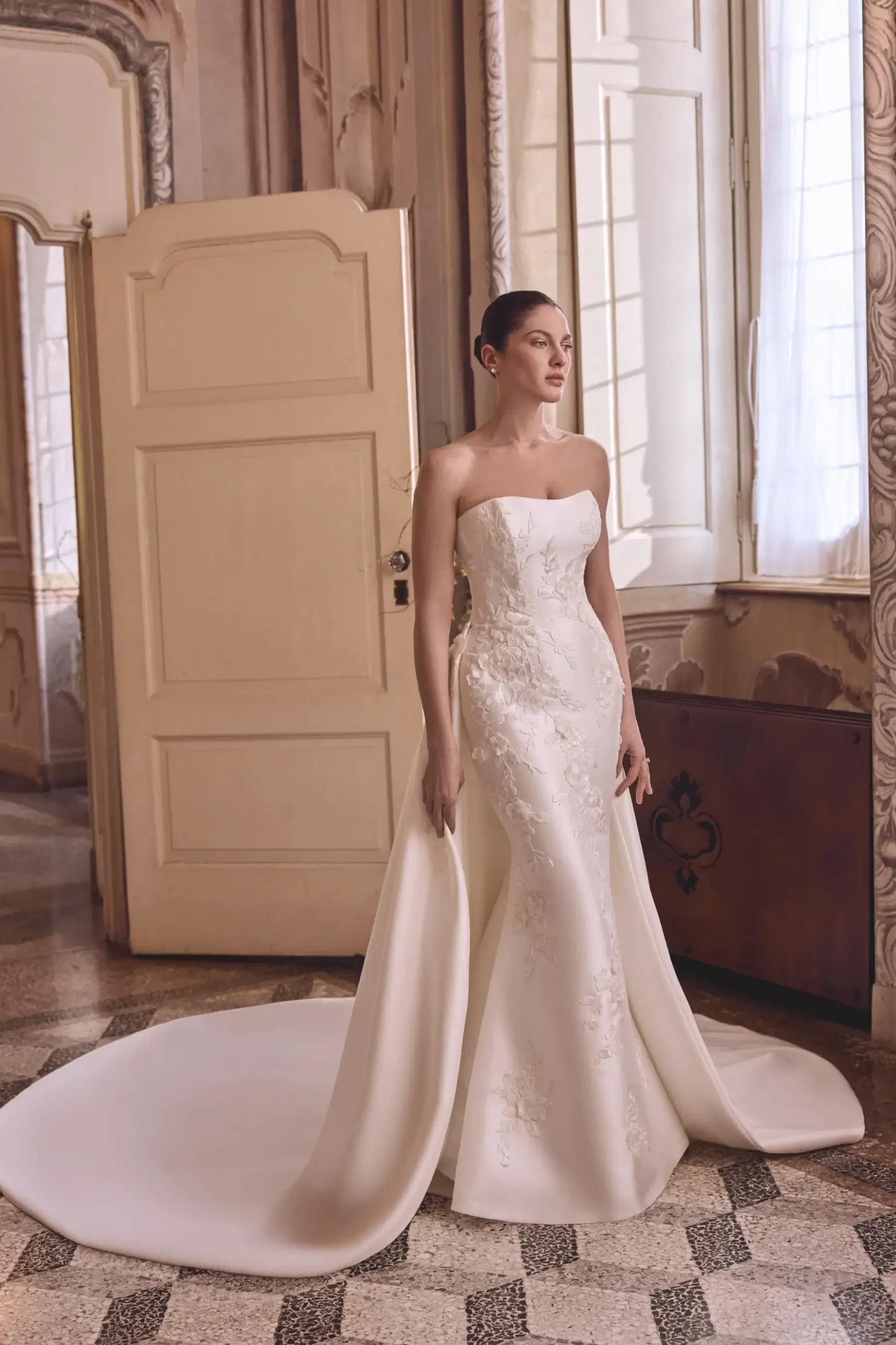 A woman in a strapless white wedding gown stands elegantly in a room with large windows and ornate decor.