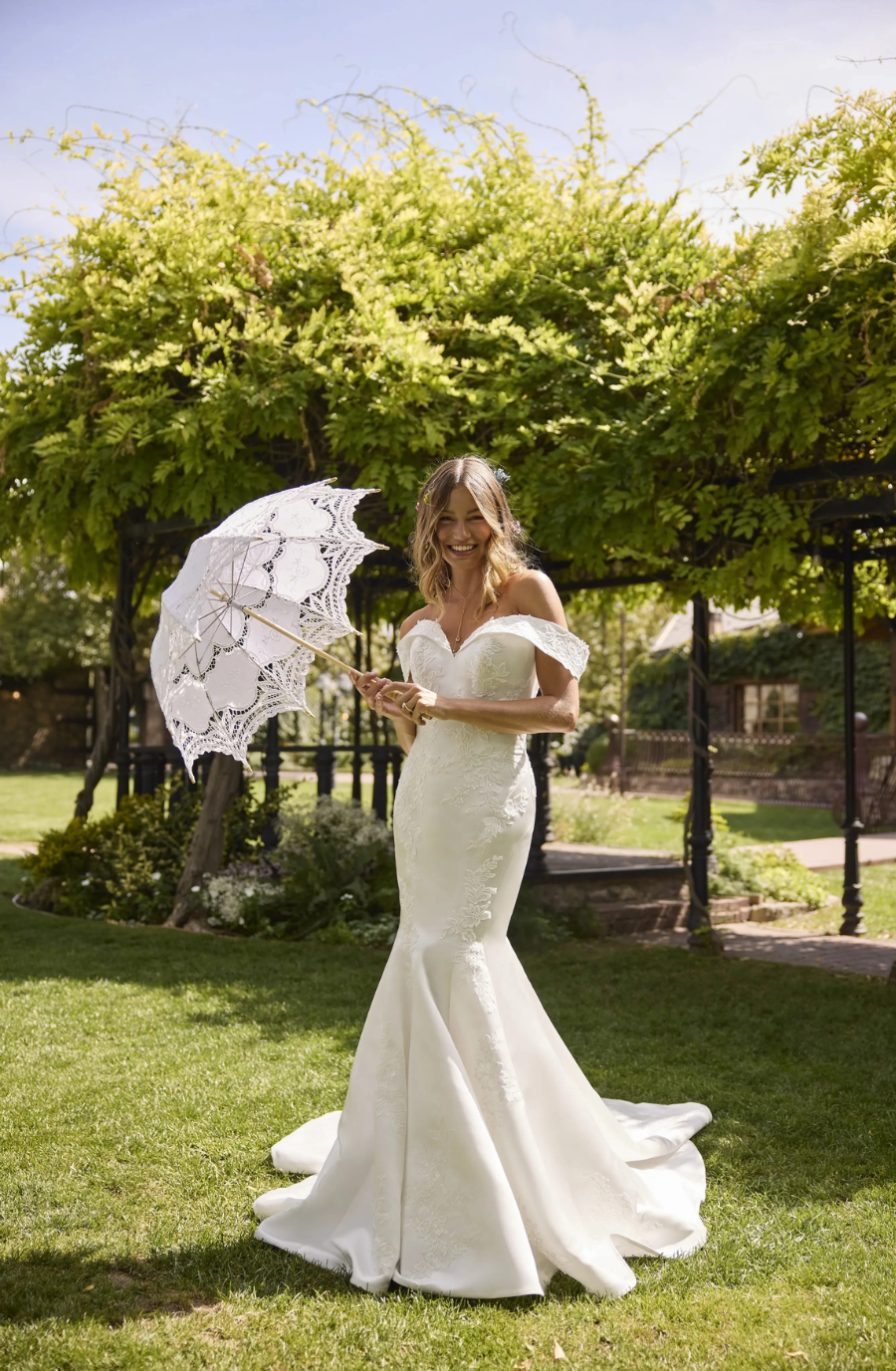 Bride in an elegant white gown stands on grass, holding a lace parasol. She is smiling under a lush, green pergola, exuding joy and serenity.