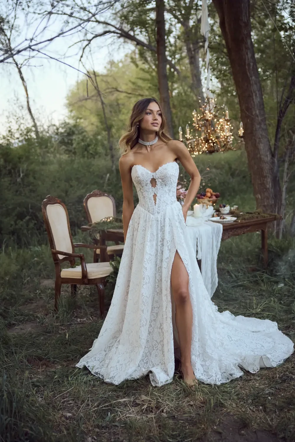 A woman wearing a white lace wedding dress with a thigh-high slit stands in a forested area, beside a table set for a meal.