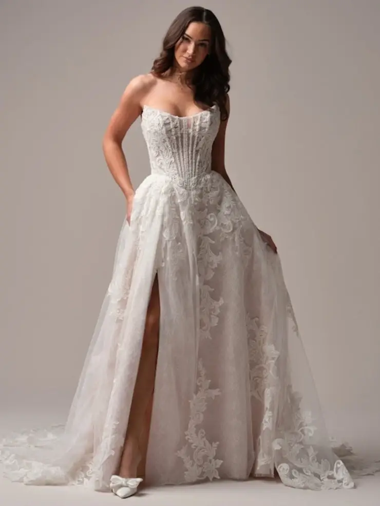 Model is wearing a strapless a-line wedding dress with a corset bodice and lace fabric with a thigh high slit