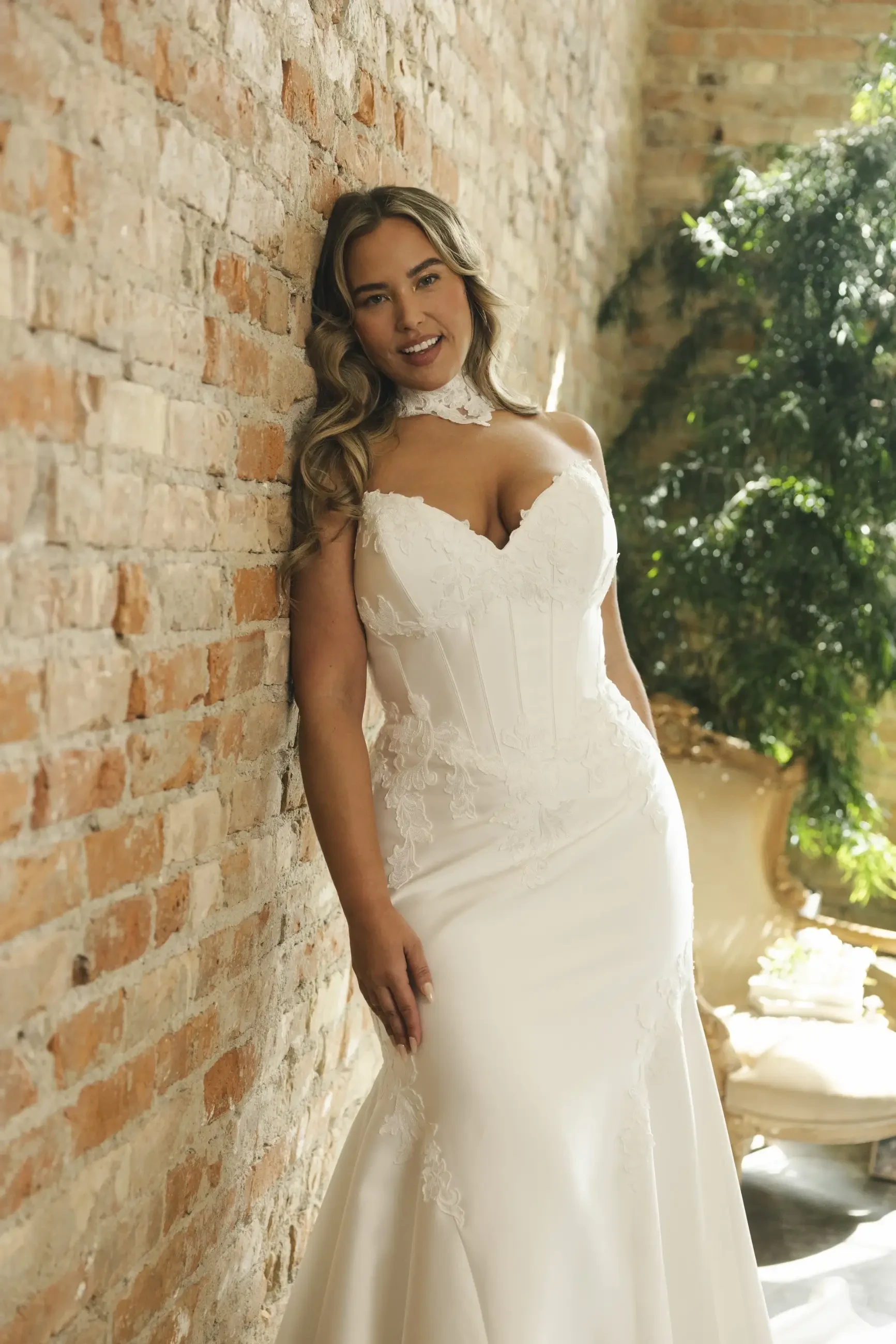 A woman in a strapless white lace wedding dress leans against a rustic brick wall, smiling. Soft sunlight filters through greenery, creating a romantic atmosphere.