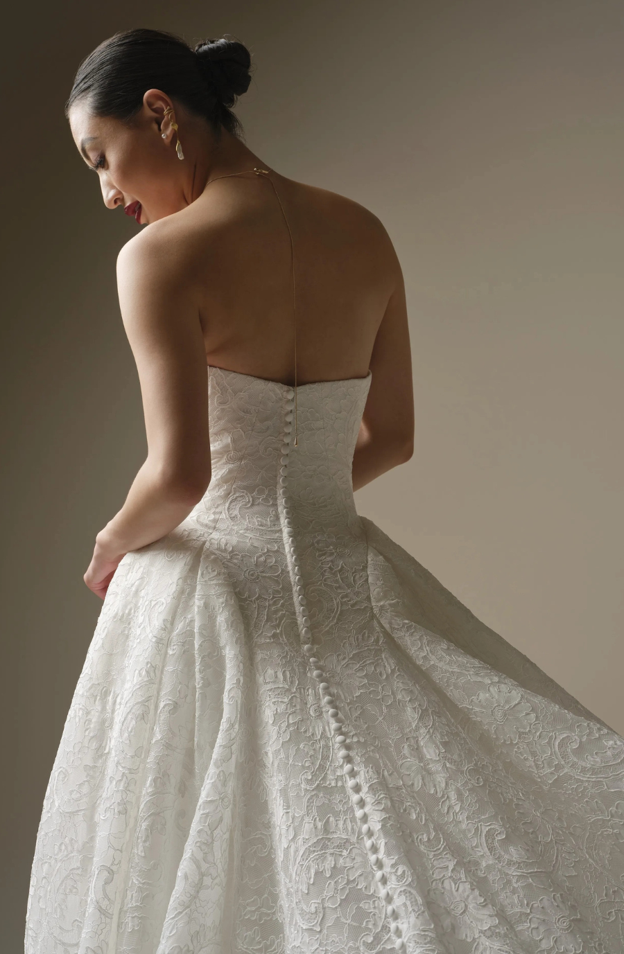 A woman in an elegant, strapless lace wedding gown with a flowing skirt stands in profile. Her hair is in a bun, conveying elegance and grace.