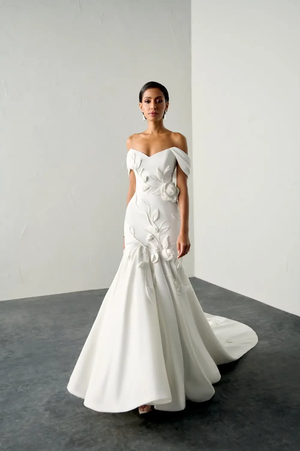A model wearing an elegant white off-the-shoulder gown with floral embellishments, standing in a minimalist space.