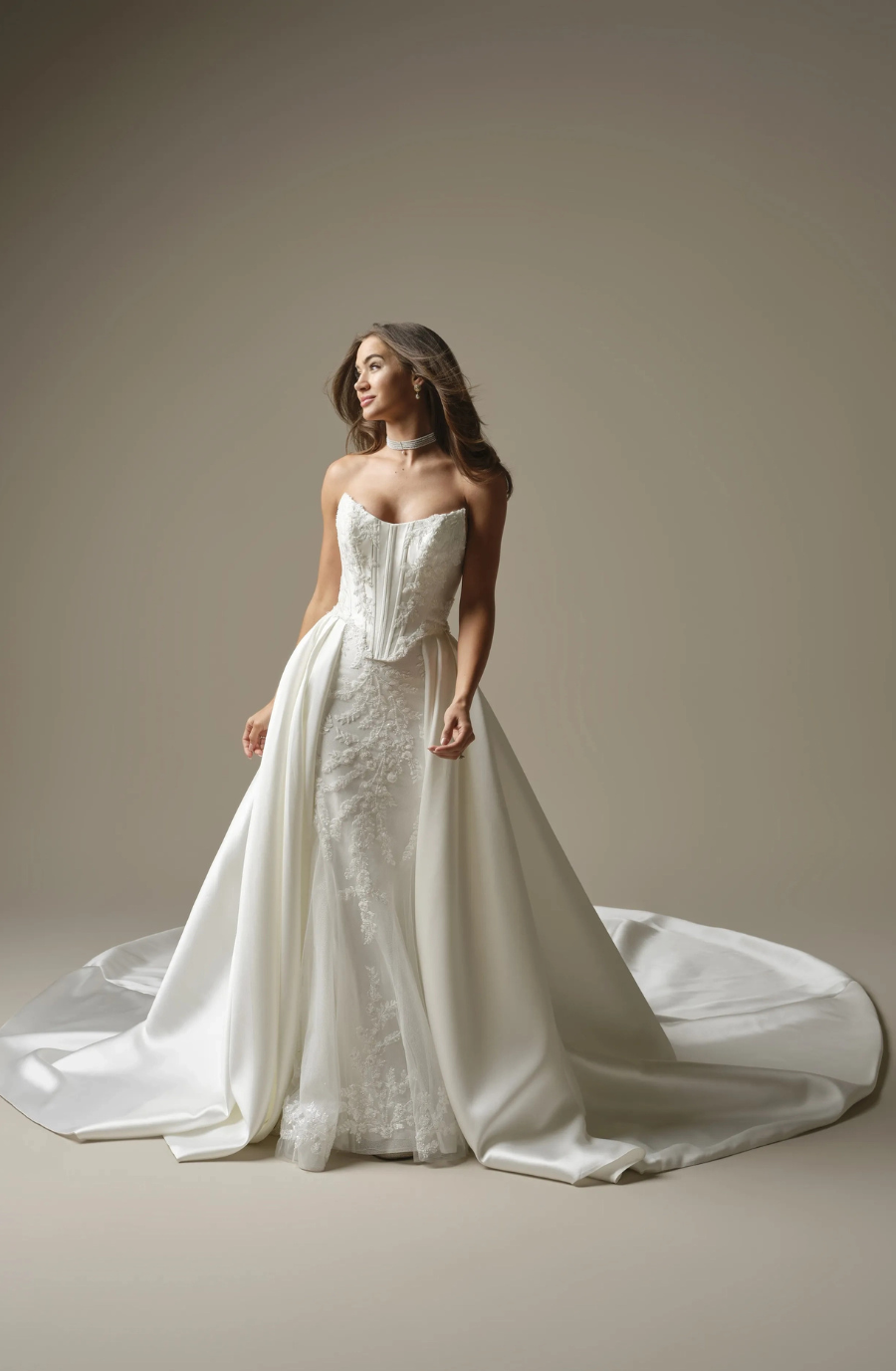 A woman in an elegant strapless white wedding dress with intricate lace and a long, flowing train stands confidently against a neutral background.